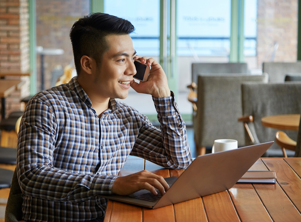 Asian man working on a laptop while on a phone call at his desk